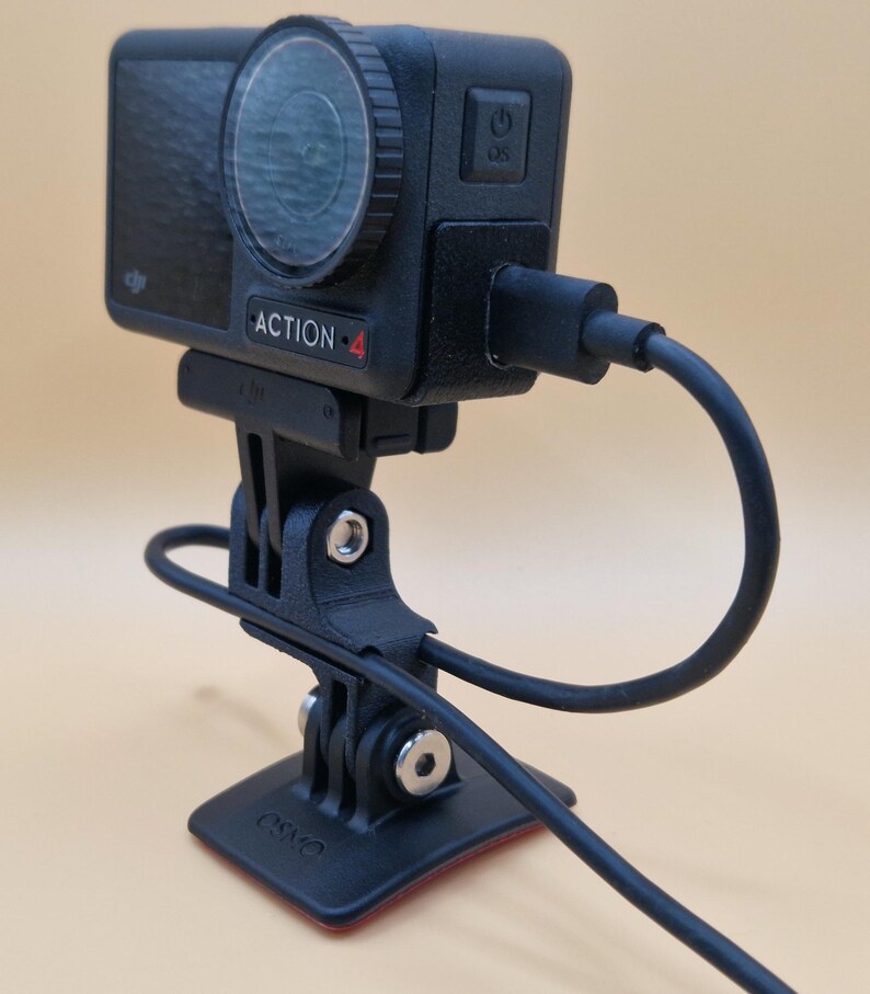 May include: Black action camera labeled "ACTION-4" with a lens and power button, mounted on a black bracket with a cable attached. The camera is connected to a black base with a red adhesive.