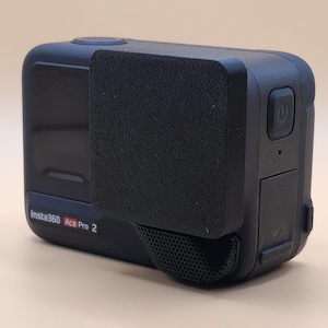 Puede incluir: Cámara de acción negra Insta360 Ace Pro 2 con una cubierta protectora negra. La cámara tiene una pequeña pantalla lateral y un botón de encendido en el lado derecho.