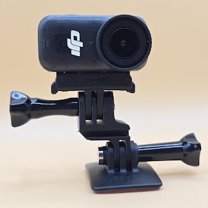 May include: A black DJI Osmo action camera mounted on a black plastic adjustable bracket system. The camera features a prominent circular lens and the DJI logo on the front. The mounting base includes a flat square plate with a thumb screw.