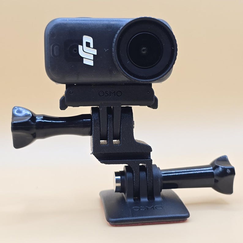 May include: A black DJI Osmo action camera mounted on a black plastic adjustable bracket system. The camera features a prominent circular lens and the DJI logo on the front. The mounting base includes a flat square plate with a thumb screw.