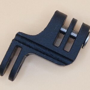 May include: A black, angled bicycle accessory with a slotted design. It features two parallel slots on one end and three on the other, with silver screws visible. The accessory is likely for mounting or securing equipment on a bicycle.