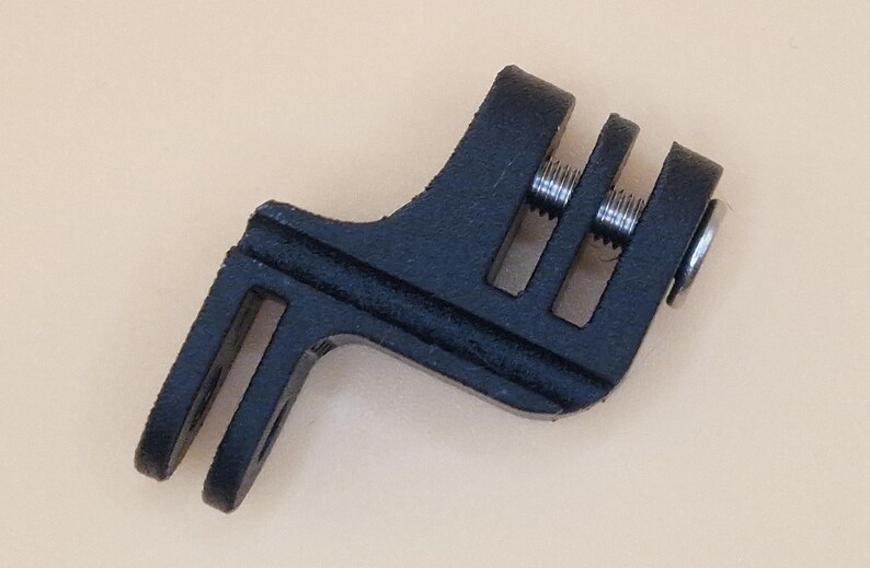 May include: A black, angled bicycle accessory with a slotted design. It features two parallel slots on one end and three on the other, with silver screws visible. The accessory is likely for mounting or securing equipment on a bicycle.
