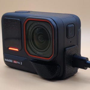 Può includere: Una fotocamera Insta360 One RS nera con un bordo rosso attorno all'obiettivo. La fotocamera è collegata a un cavo di ricarica nero. La fotocamera ha un piccolo schermo laterale e un microfono nella parte inferiore.