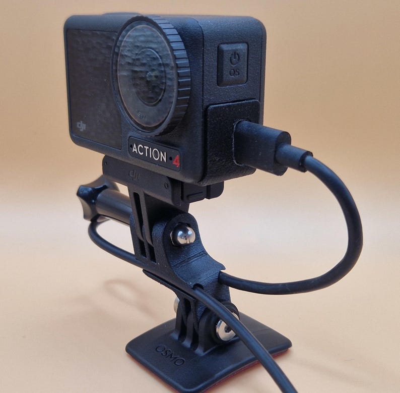May include: Black action camera with the text "ACTION 4" on the front, mounted on a black stand. The camera has a lens and a power button. A black cable is plugged into the camera. The stand has a base with the text "OSMO".