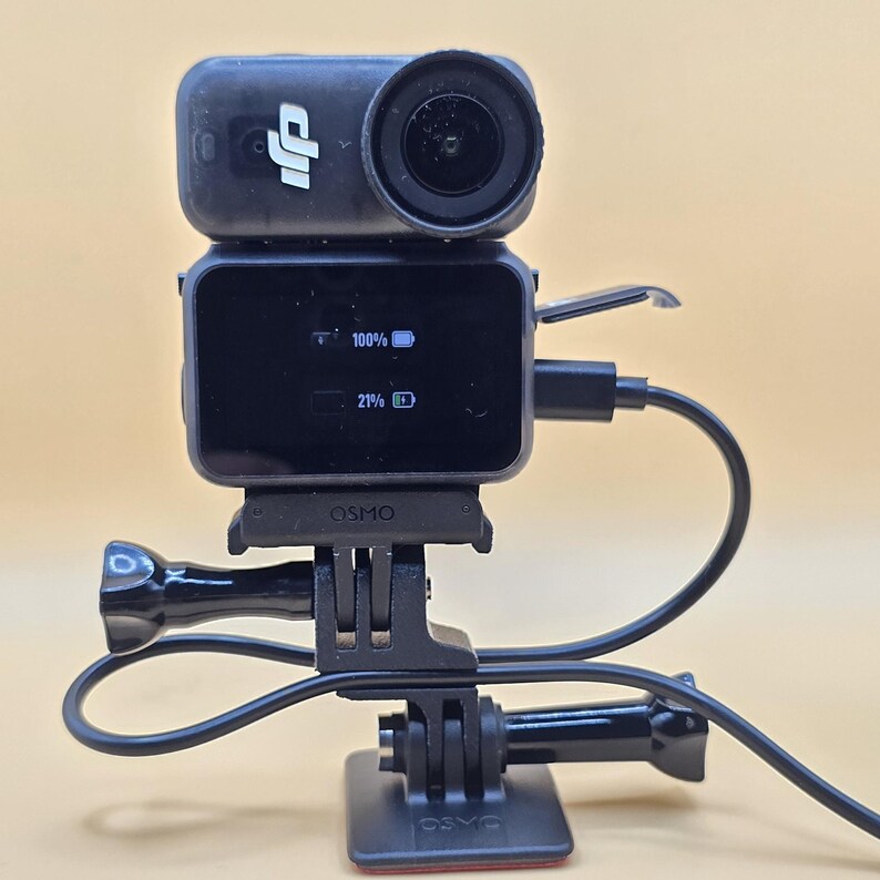 May include: A black DJI Osmo action camera mounted on a black plastic base. The screen displays two battery icons with 100% and 21% charge levels. A black charging cable is plugged into the side of the camera, and the word Osmo is visible on the mount.