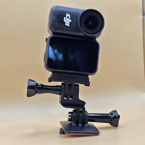 May include: A black DJI Osmo action camera mounted on a black plastic adjustable tripod adapter. The camera features a prominent circular lens on the front and a rectangular display screen. The word Osmo is embossed on the camera body and the base.