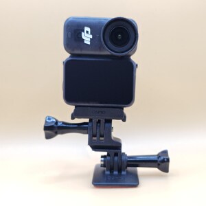 May include: A black DJI Osmo action camera mounted on a black plastic base with an adjustable hinge. The camera features a circular lens on the right, a small DJI logo on the left, and a rectangular screen on the front. The word Osmo is embossed.