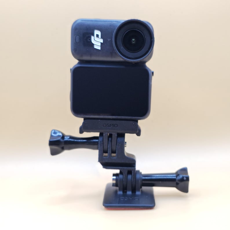 May include: A black DJI Osmo action camera mounted on a black plastic base with an adjustable hinge. The camera features a circular lens on the right, a small DJI logo on the left, and a rectangular screen on the front. The word Osmo is embossed.