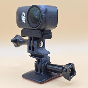May include: A black DJI Osmo action camera mounted on a black plastic adjustable bracket with a base plate. The camera features a circular lens and the DJI logo on the front. The mounting system includes a screw knob and a flat adhesive base.