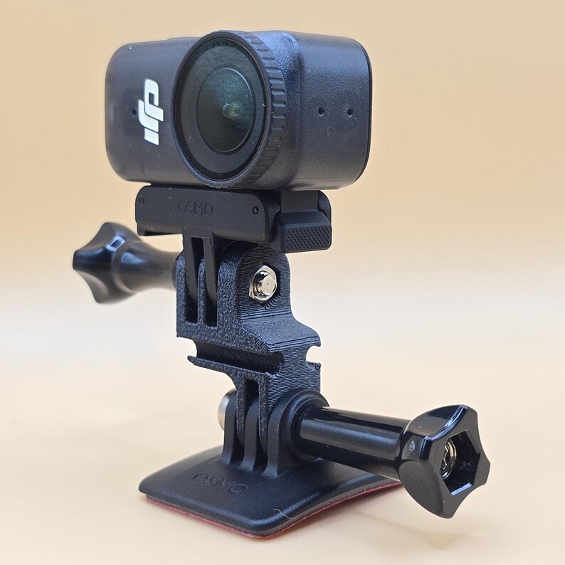 May include: A black DJI Osmo action camera mounted on a black plastic adjustable bracket with a base plate. The camera features a circular lens and the DJI logo on the front. The mounting system includes a screw knob and a flat adhesive base.