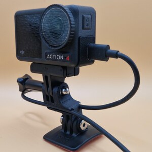 May include: Black Action 4 camera with a lens, mounted on a black bracket and base. The camera is connected to a black cable. The camera has the word "ACTION 4" printed on it.