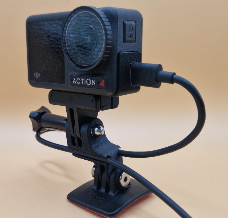 May include: Black Action 4 camera with a lens, mounted on a black bracket and base. The camera is connected to a black cable. The camera has the word "ACTION 4" printed on it.