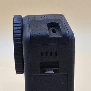 Puede incluir: Una cámara de acción negra con una carcasa de lente circular con un borde estriado en el lateral. El cuerpo del dispositivo tiene una cubierta de puerto rectangular en la parte superior, cuatro pequeñas ranuras de ventilación y un pestillo.