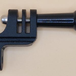 May include: A black camera mount adapter with a ball joint, screw, and a rectangular base with two slots. The adapter has a cylindrical extension with a curved end. The item is designed to attach to a camera.