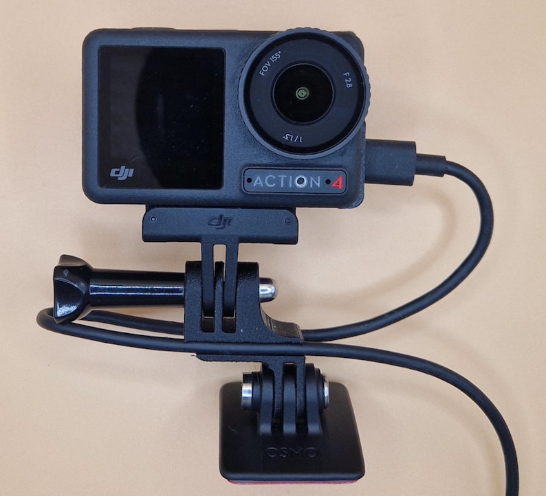 May include: Black DJI Action 4 camera with a lens, screen, and attached mount. The camera is connected to a black mount and base with a black cable. The camera is mounted on a beige surface.