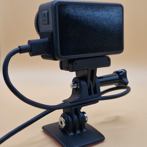 May include: A black action camera with a screen, mounted on a black tripod. The camera is connected to a black cable. The tripod has a base with an adhesive pad.