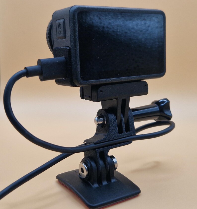 May include: A black action camera with a screen, mounted on a black tripod. The camera is connected to a black cable. The tripod has a base with an adhesive pad.