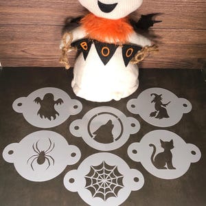 May include: A white ghost doll with black eyes and a smiling mouth, wearing an orange collar and holding a banner that says "BOO". Includes Halloween-themed stencils with a ghost, spider, witch, cat, wolf, and spiderweb designs.