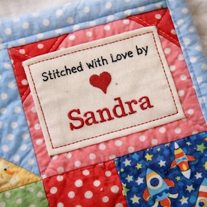 May include: Close-up of a handmade quilt with a patchwork design. The quilt features a label that reads "Stitched with Love by Sandra" with a red heart. The quilt has blue polka dot, red polka dot, and rocket ship fabric.