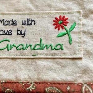 May include: A rectangular fabric label stitched onto a quilt, featuring the embroidered black text Made with love by and the green text Grandma, accompanied by a small red flower with green leaves, set against a beige and floral fabric background.