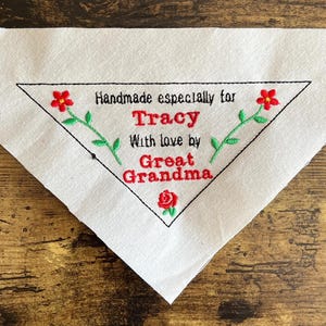 May include: A white triangular fabric patch featuring black embroidered text that reads Handmade especially for Tracy With love by Great Grandma. The design includes red flower accents with green stems and leaves on the sides and bottom.