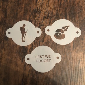 May include: Three white stencils with brown designs and text. One stencil features a soldier with a rifle, another a poppy flower, and the third reads "Lest We Forget". The stencils have a circular shape with two small side tabs.