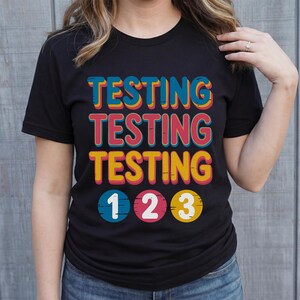 Puede incluir: Camiseta negra con la palabra "TESTING" repetida tres veces en una tipograf&iacute;a retro colorida. Debajo, los n&uacute;meros "1 2 3" en c&iacute;rculos. Una prenda casual para el d&iacute;a a d&iacute;a.