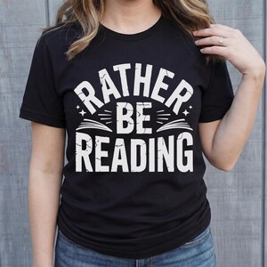 May include: Black t-shirt with the white text "RATHER BE READING". The text is arched above a graphic of an open book with stars. The shirt is a crew neck and is being worn by a person.