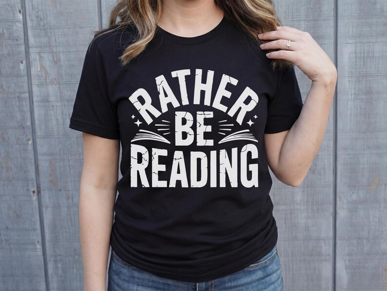 May include: Black t-shirt with the white text "RATHER BE READING". The text is arched above a graphic of an open book with stars. The shirt is a crew neck and is being worn by a person.