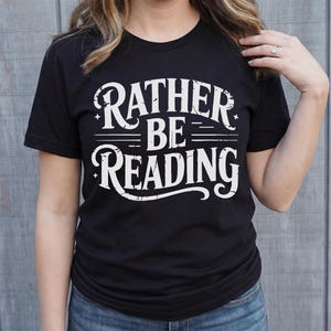 May include: Black t-shirt with the white text "RATHER BE READING" in a distressed, vintage style. The shirt is a crew neck and is being worn by a person. The background is a light gray wooden wall.