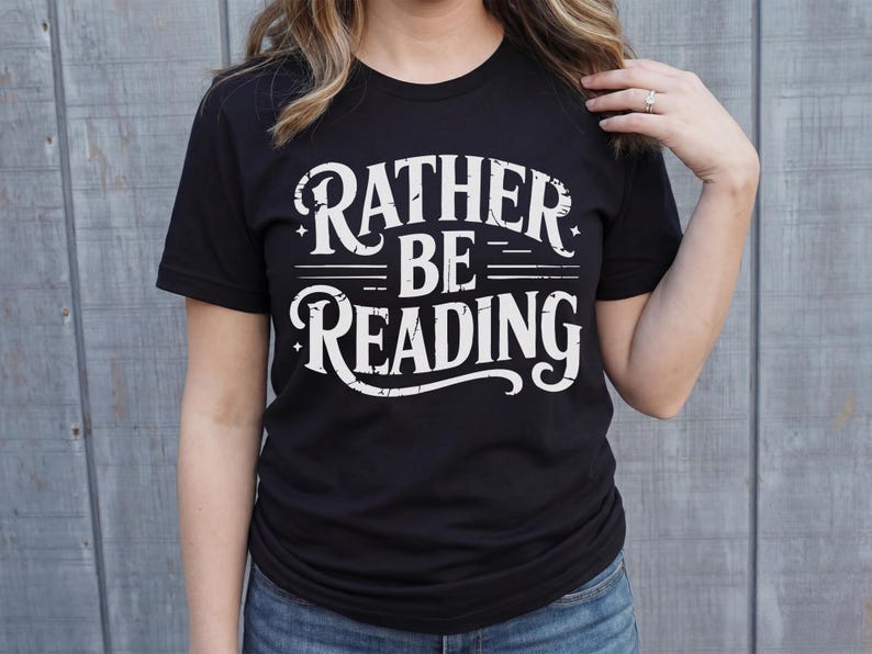 May include: Black t-shirt with the white text "RATHER BE READING" in a distressed, vintage style. The shirt is a crew neck and is being worn by a person. The background is a light gray wooden wall.