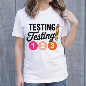 Puede incluir: Camiseta blanca con las palabras "TESTING Testing" en negro, un dibujo de l&aacute;piz amarillo y los n&uacute;meros 1, 2 y 3 en rosa, melocot&oacute;n y naranja. La camiseta la lleva una persona.