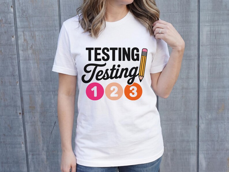 Puede incluir: Camiseta blanca con las palabras "TESTING Testing" en negro, un dibujo de l&aacute;piz amarillo y los n&uacute;meros 1, 2 y 3 en rosa, melocot&oacute;n y naranja. La camiseta la lleva una persona.