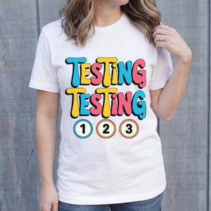 Puede incluir: Camiseta blanca con las palabras "TESTING TESTING" en letras coloridas de estilo retro. Debajo del texto hay tres c&iacute;rculos numerados: 1, 2 y 3. La camiseta est&aacute; siendo usada.
