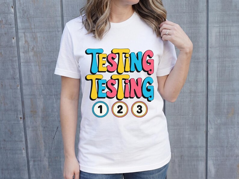 Puede incluir: Camiseta blanca con las palabras "TESTING TESTING" en letras coloridas de estilo retro. Debajo del texto hay tres c&iacute;rculos numerados: 1, 2 y 3. La camiseta est&aacute; siendo usada.