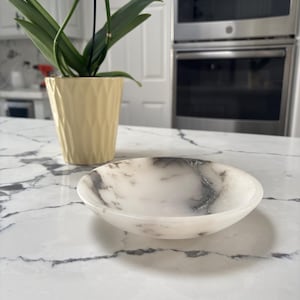 May include: A shallow, circular bowl crafted from white stone with prominent grey veining throughout its surface. The bowl features a smooth, polished finish and a curved rim, presenting a natural marbled appearance with distinct dark grey streaks.