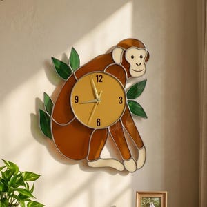 May include: A decorative monkey-shaped wall clock with a brown body, cream-colored face and hands, and a yellow clock face. The clock is surrounded by green leaves. The clock is mounted on a beige wall.