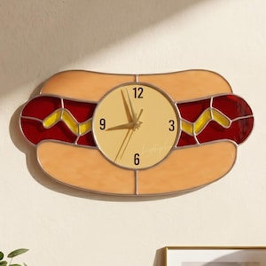 May include: A novelty wall clock shaped like a hot dog. The clock face is round with black numerals and hands. The hot dog design features a tan bun, red and yellow accents, and the text "LightupLux" in the upper left corner.