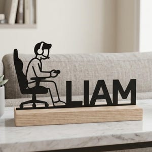Peut inclure: Figurine de gamer en métal noir avec le nom "LIAM" sur une base en bois. La figurine représente une personne dans un fauteuil de jeu portant un casque et tenant une manette. Le texte "Lightuplux" est dans le coin supérieur gauche.