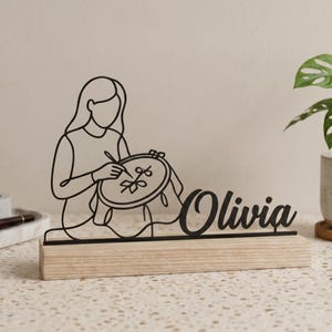 May include: Black metal desk ornament depicting a person embroidering, with the name "Olivia" in cursive. The design is mounted on a light wood base. A small plant in a white pot is to the right.