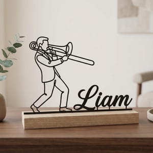 Op de afbeelding: Zwart metalen silhouet van een trombonist op een houten voet met de naam "Liam" in cursief. Het kunstwerk staat op een donkere houten tafel, met een vaas en groen links.