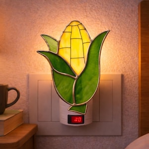 May include: A stained glass night light shaped like an ear of corn, with yellow kernels and green leaves. The night light is plugged into a wall outlet and has a small digital display. The brand name "LightupLux" is visible.