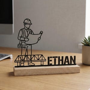 May include: A black metal desk nameplate featuring an engineer figure holding blueprints, standing on a bridge design, with the name "ETHAN". The nameplate is mounted on a wooden base. The "Lightuplux" logo is in the upper left corner.