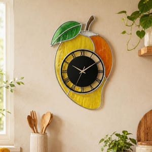 May include: A mango-shaped wall clock with a black clock face and Roman numerals. The clock features a yellow and orange stained glass design with a green leaf and brown stem. The clock is mounted on a light-colored wall.