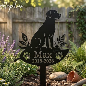 Può includere: Paletto da giardino in metallo nero con la silhouette di un cane, seduto su una placca a forma di foglia. La placca è incisa con il nome "Max" e gli anni "2018-2026". Il paletto è in un giardino con fiori e una staccionata di legno sullo sfondo.