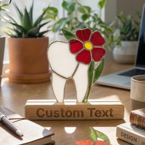 May include: A stained glass tooth with a red and yellow flower design. The tooth is white with a black outline, and the flower has red petals and a yellow center. The base is a wooden block with the words "Custom Text".