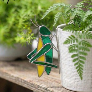 Pode incluir: Um gafanhoto de vitral verde e amarelo, com uma moldura de metal prateado, está preso a um vaso de cerâmica branco. O gafanhoto está perto de plantas verdes, com o texto "LightupLux" no canto superior esquerdo. Decoração de jardim.
