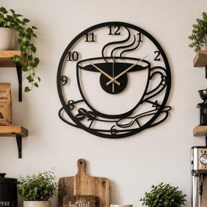 May include: Black metal wall clock with a coffee cup design. The clock features a cup, saucer, and spoon silhouette, with the numbers 1 through 12 around the edge. The clock hands are gold. The words "but first coffee" are on a wooden sign.