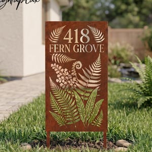 May include: A rectangular, rusty-brown metal address sign with the text "418 FERN GROVE" in silver. The sign features a fern leaf design and is mounted on two metal stakes, standing in a grassy yard. The brand name "LightupLux" is in the top left corner.