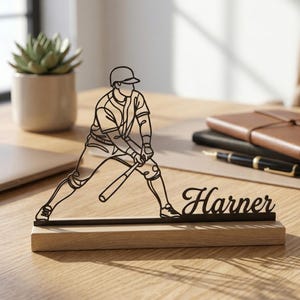 Könnte beinhalten: Schwarze Metall-Silhouette eines Baseballspielers auf einem Holzsockel, mit dem Namen "Harner" in Schreibschrift. Das Kunstwerk steht auf einem Schreibtisch mit einer Sukkulente, Notizbüchern und einem Stift. Der Markenname "LightupLux" ist oben links.